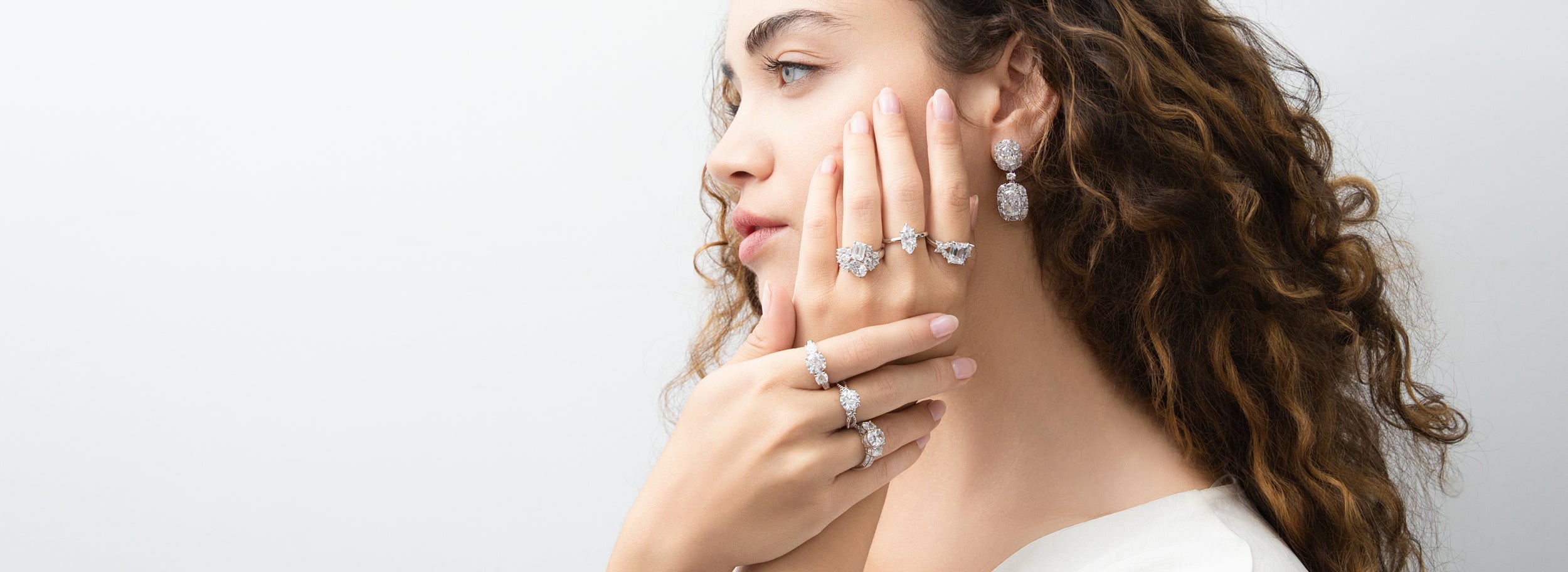 Woman with curly hair wearing multiple sparkling statement rings and drop earrings, posing against a soft white background.
