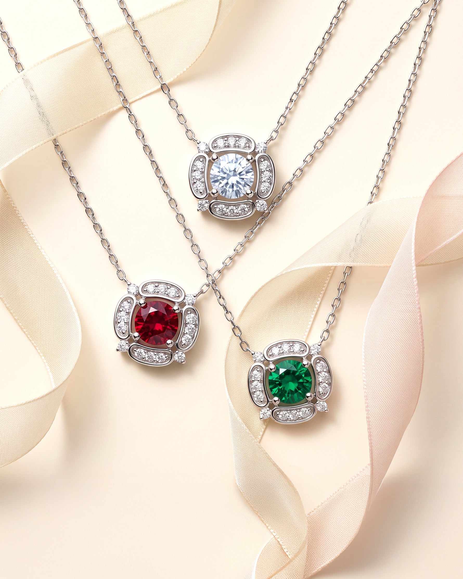 Three silver pendant necklaces with round-cut stones are arranged on a cream background with soft ribbon draping. The top pendant is clear, the middle is red, and the bottom is green, each surrounded by small clear accent stones.