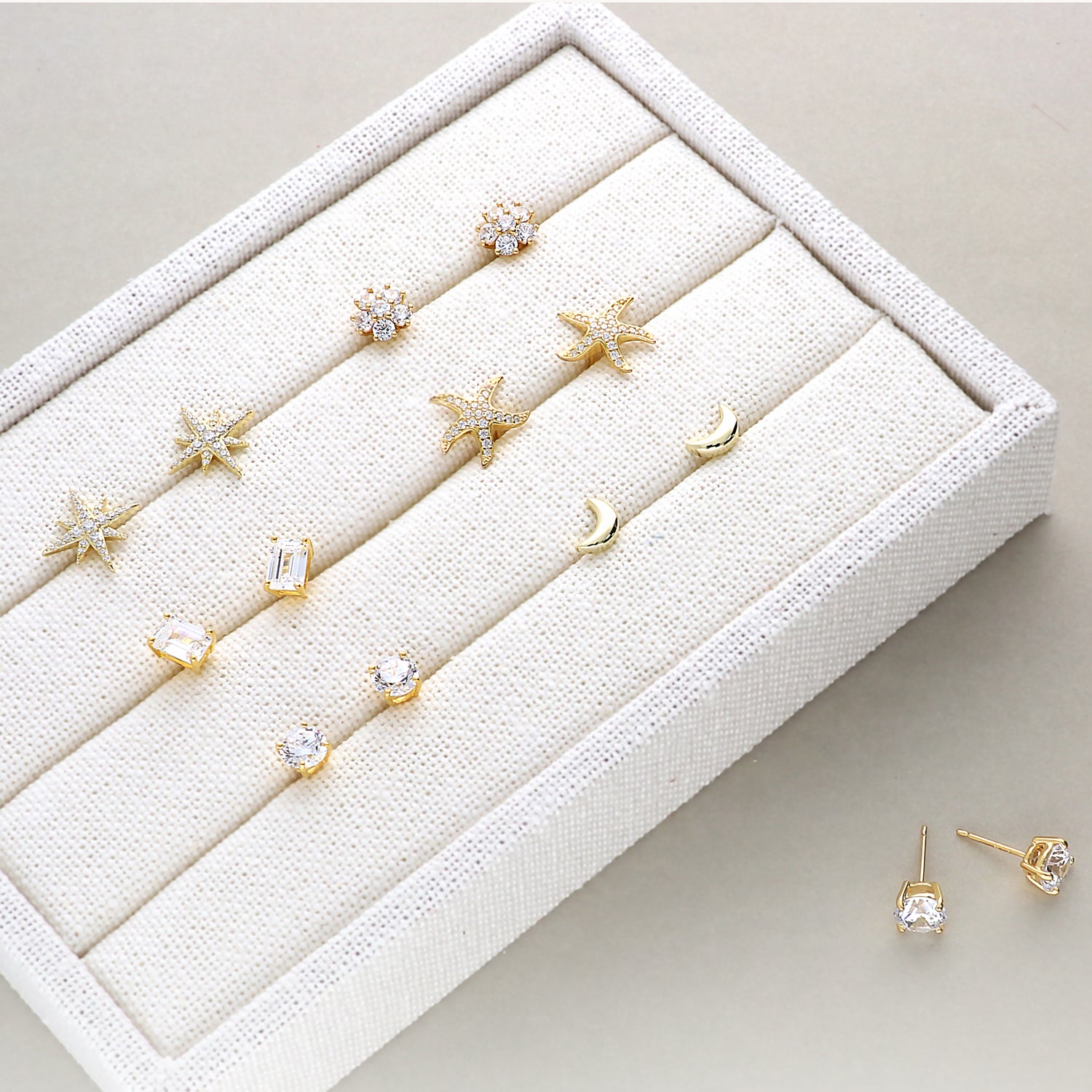 A display of various gold flashed stud earrings arranged neatly in a cream-colored jewelry box. The collection includes starfish-shaped studs, crescent moons, baguette-cut stones, and classic round-cut stone, all sparkling with elegant detailing.